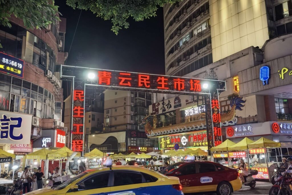 Qingyun Minsheng Market in Liuzhou at night, its lit gate sign above rows of yellow food stalls and passing taxis