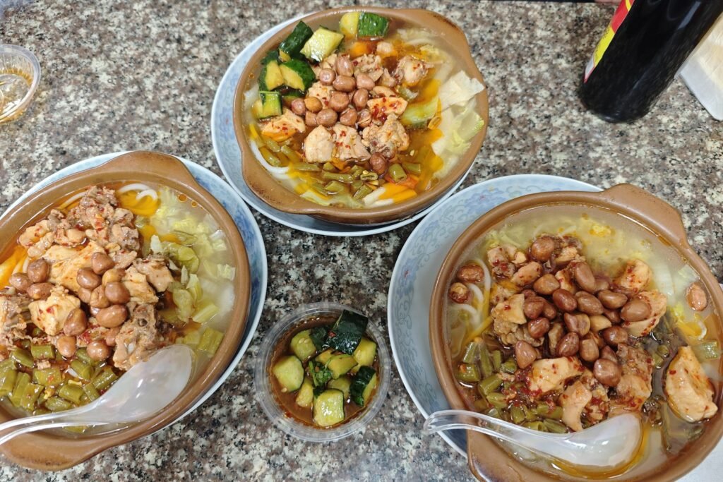 Three clay bowls of Wuchuan spicy chicken rice noodles topped with peanuts and pickled long beans, with a cup of dressed cucumber
